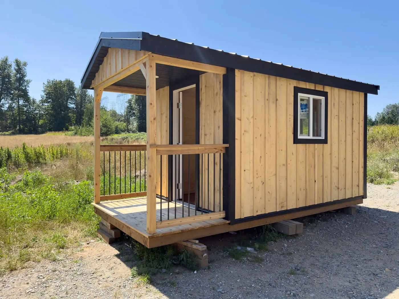 Cedar-board shed with a black metal roof and a covered porch with black railings, on a gravel pad